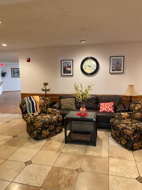 A cozy lobby seating area with a sofa, armchairs, and a table adorned with flowers, under a wall clock and framed pictures.
