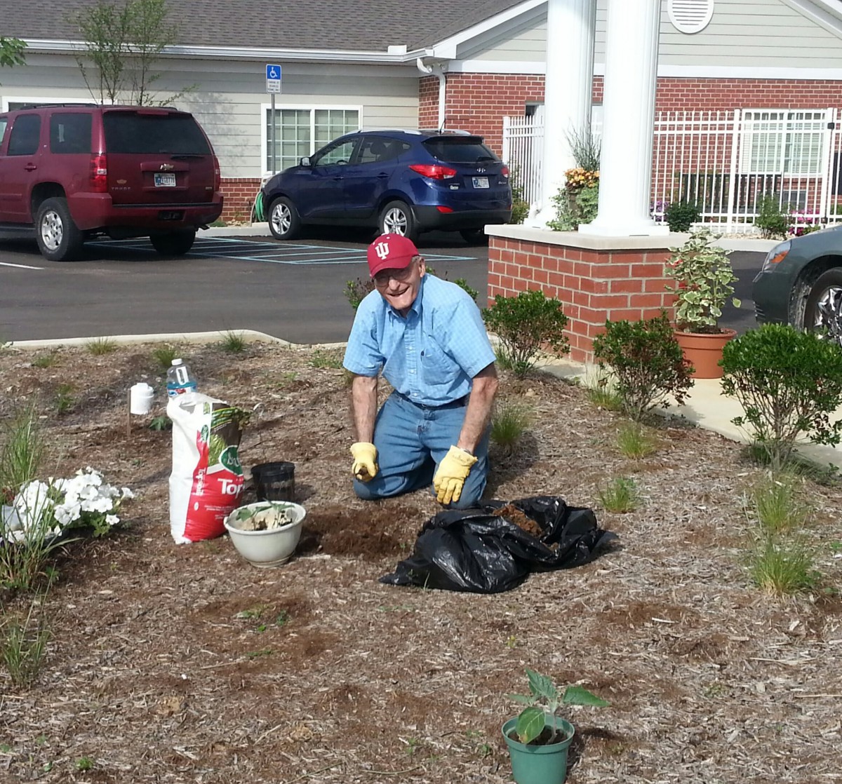 A man is gardening outside a building, surrounded by plants and tools.