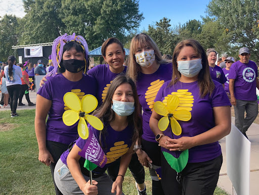 A cheerful group of participants in purple shirts holding flower props, gathered at an outdoor event.