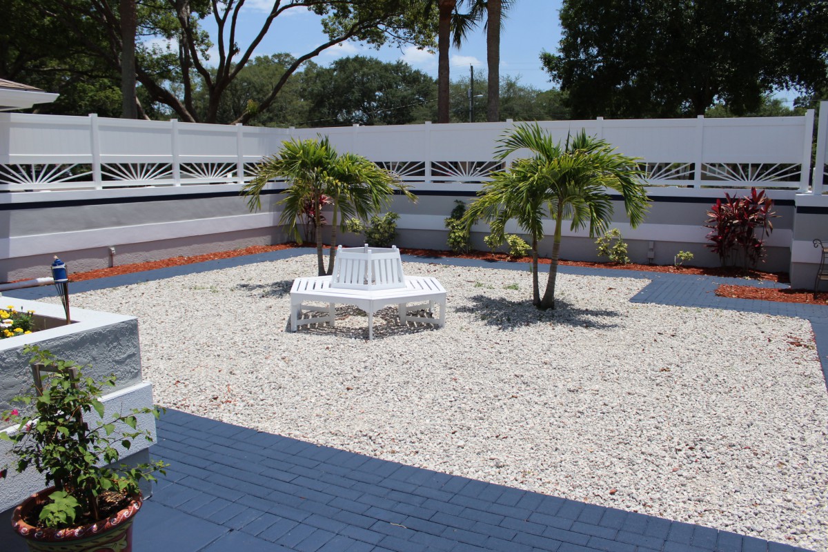 A peaceful garden area with a gravel surface, featuring palm trees and a white bench.