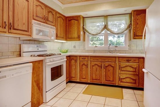 A classic kitchen with wooden cabinets, featuring a stove, oven, and microwave, accented by tiled flooring and a window with curtains.