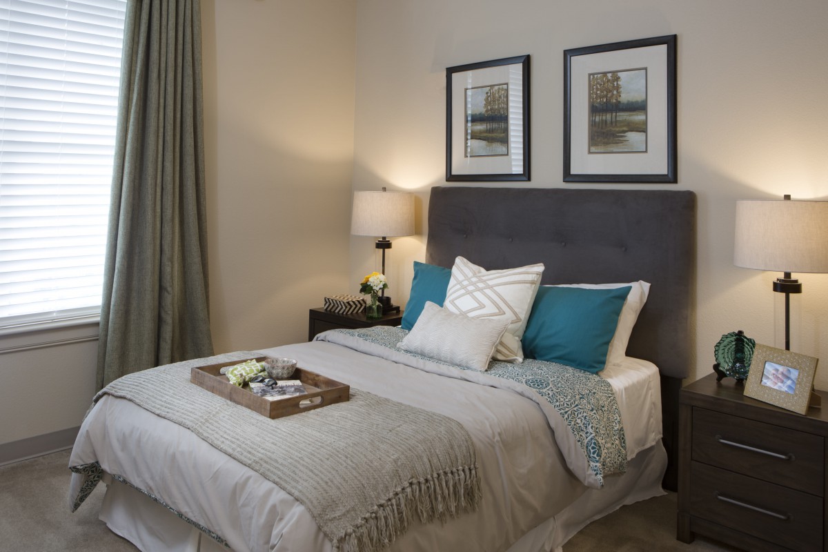 A cozy, modern bedroom with a neatly made bed featuring blue and white pillows, framed art above the headboard, and a bedside lamp.