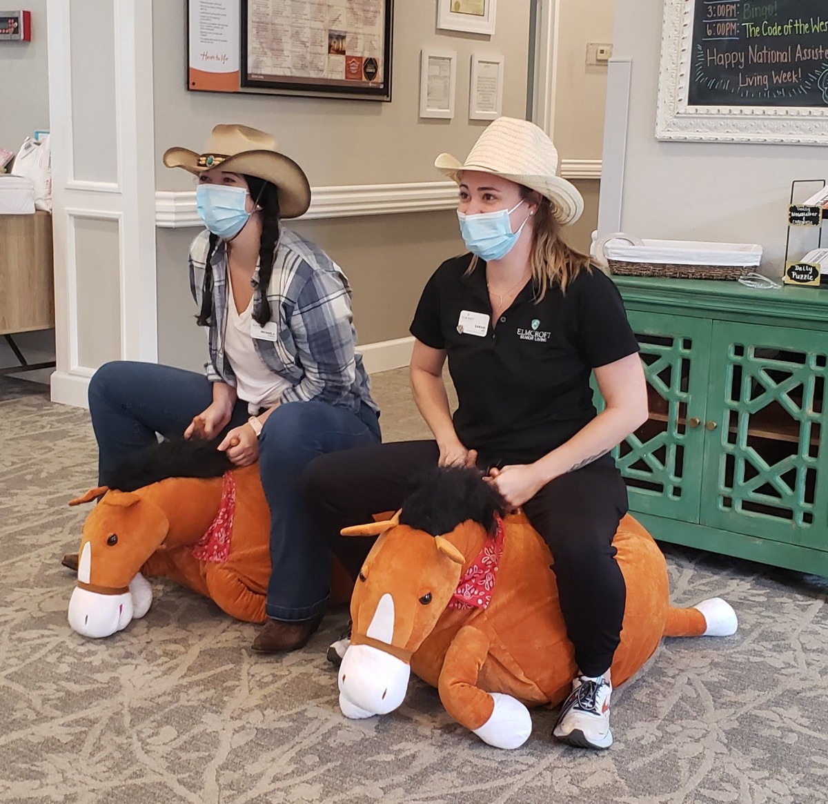 Two people wearing cowboy hats and masks sit on plush horses indoors.