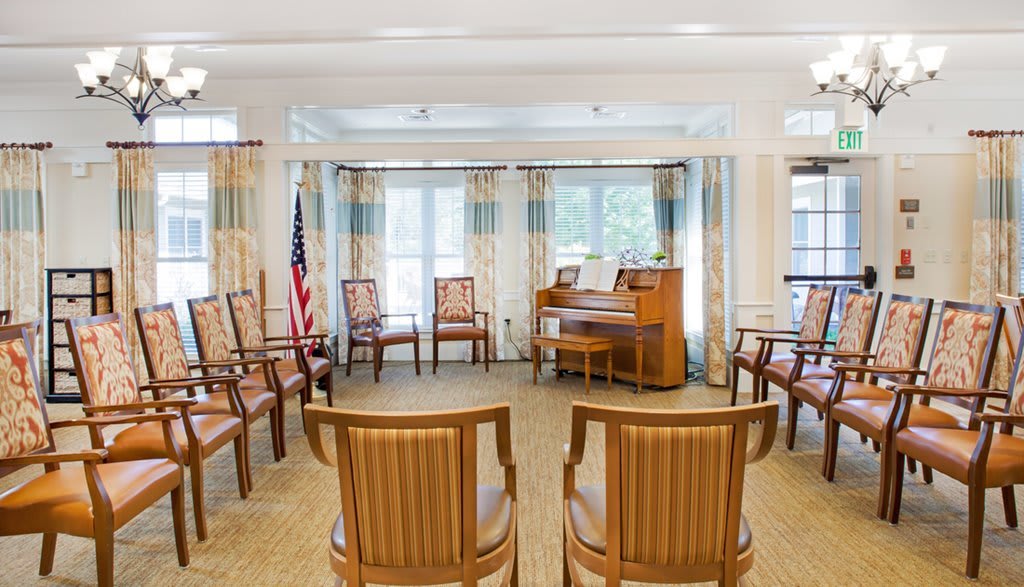 A well-lit room with arranged chairs, a piano, and large windows accompanied by curtains.