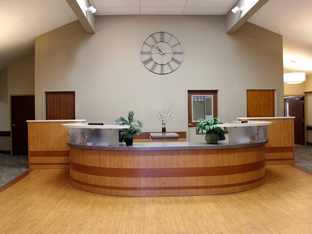 A modern office reception area with a central desk, decorative plants, and a large wall clock.