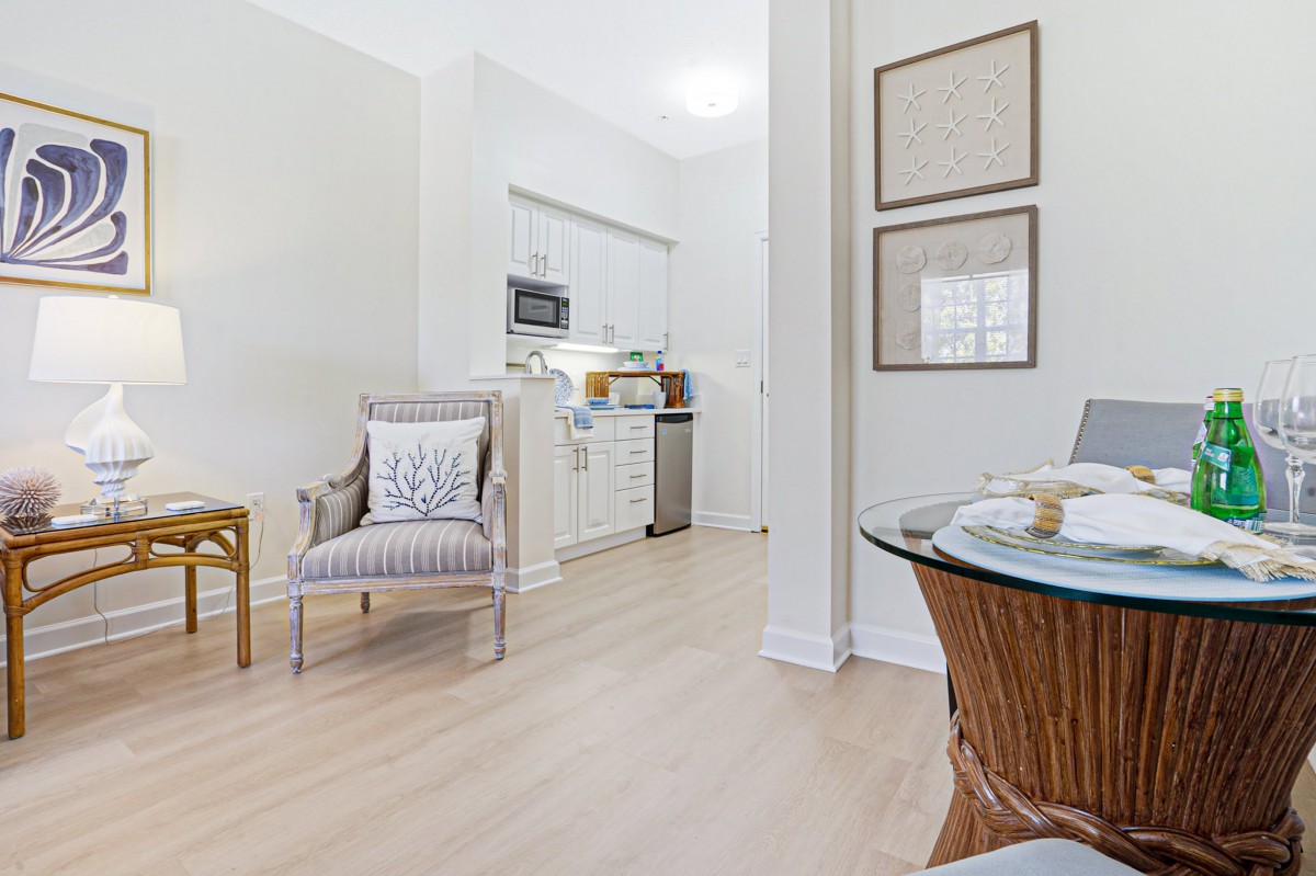 A modern open-plan living space featuring a cozy armchair, dining table, and a compact kitchen with white cabinetry.