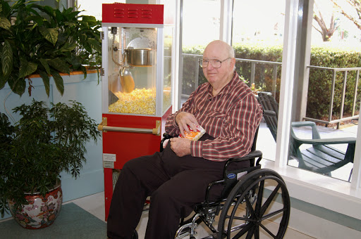 An elderly man enjoys popcorn while seated in a wheelchair next to a popcorn machine.