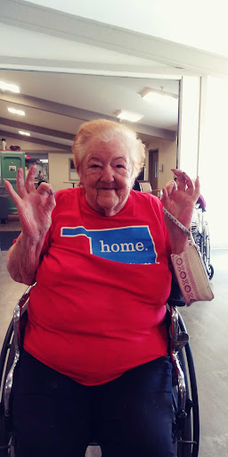 An elderly woman in a red shirt with the word 'home' and an outline of Oklahoma, smiling and giving a thumbs-up from her wheelchair.