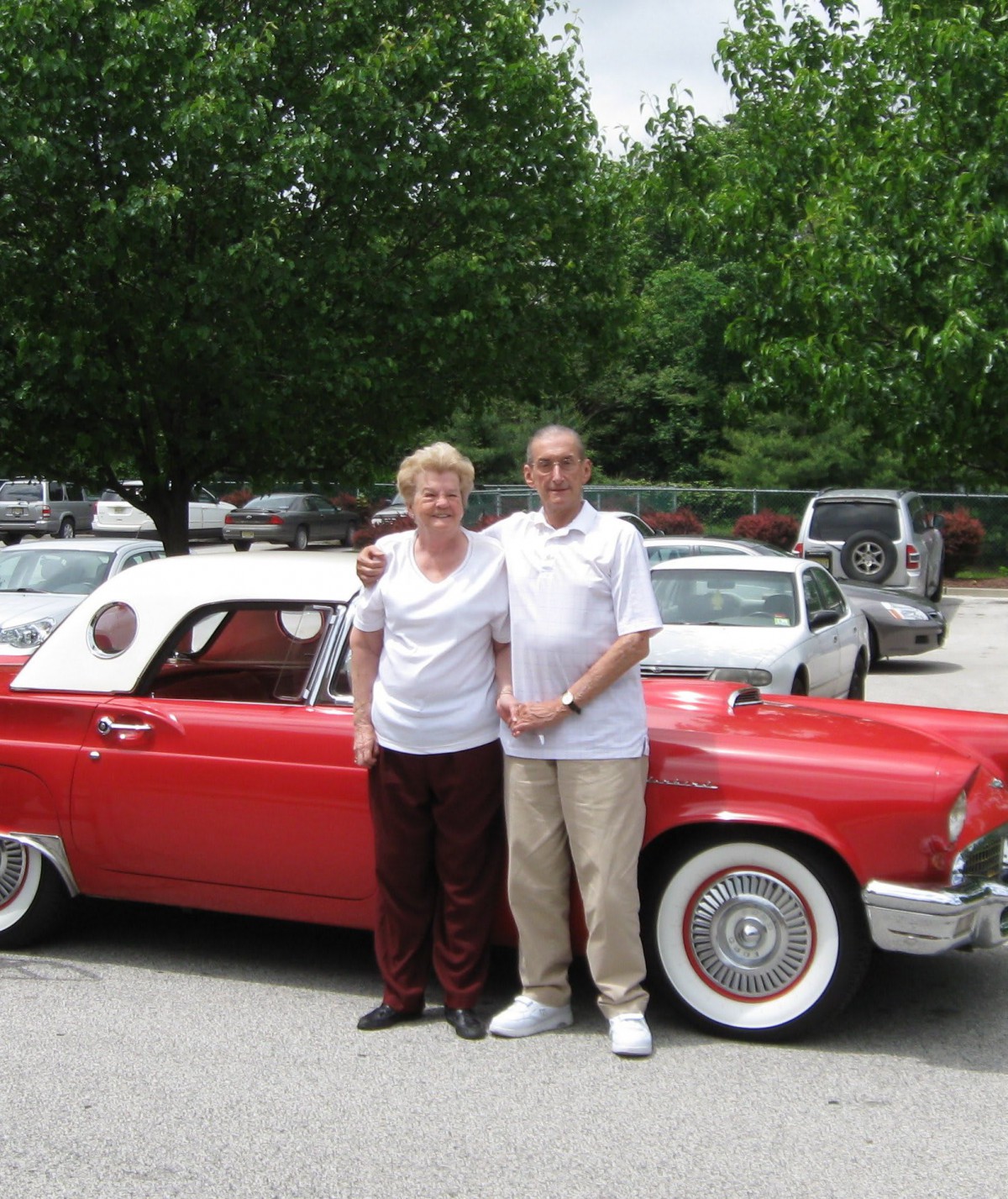 A couple stands next to a red vintage convertible in a parking lot.