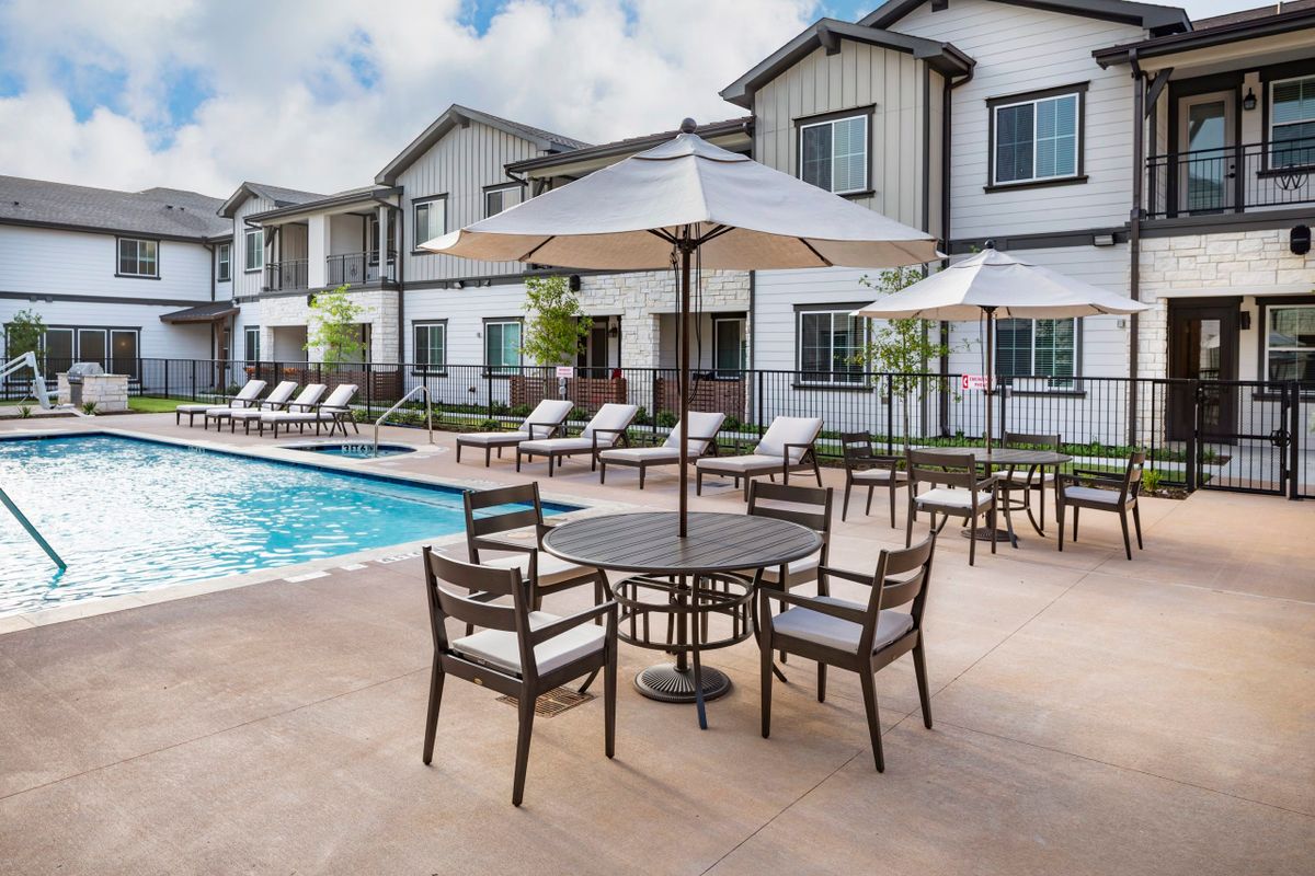 A sunny outdoor pool area with lounge chairs, tables, and umbrellas in a resort setting.