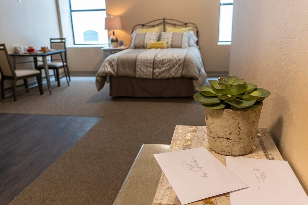A cozy bedroom featuring a neatly made bed, a small desk with a lamp, and a succulent plant on the table.