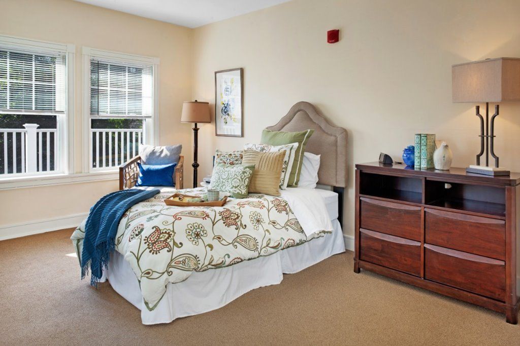 A cozy bedroom with a neatly made bed, decorative pillows, a dresser, and a window letting in natural light.