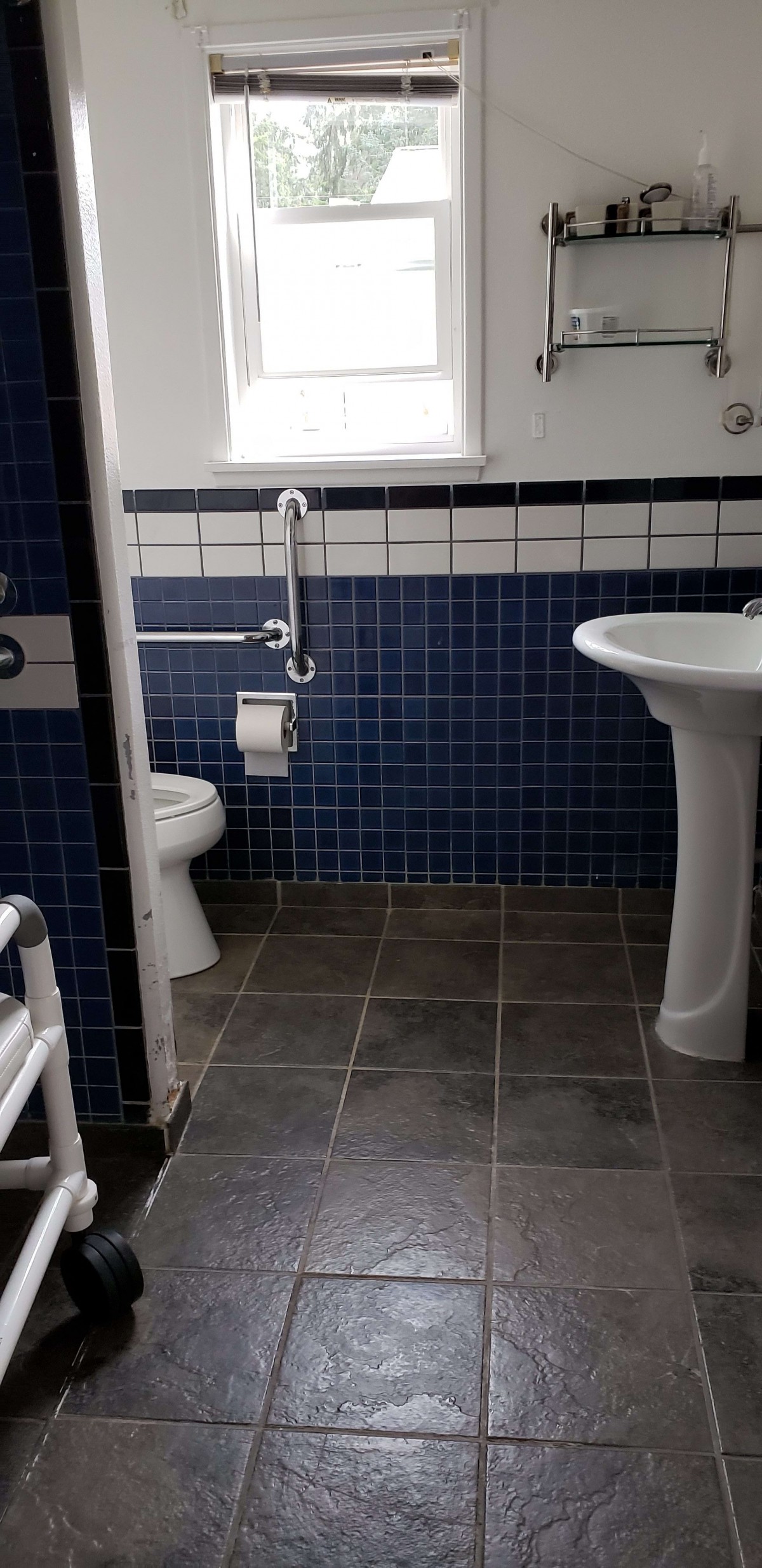 A bathroom with blue tiled walls, a pedestal sink, and a toilet with a handrail.