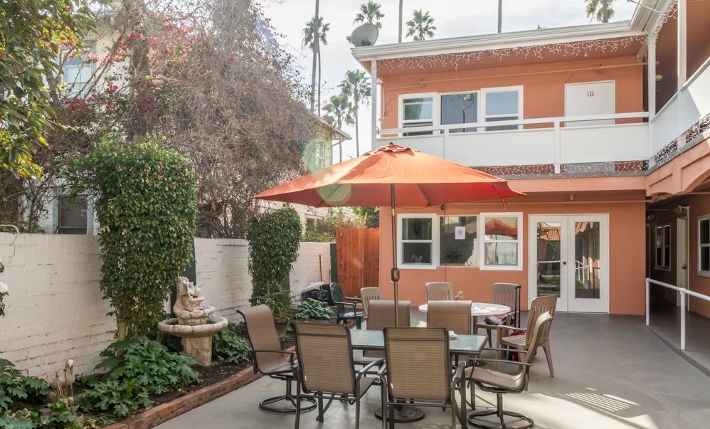 A cozy outdoor patio area with a table, chairs, and an orange umbrella in front of a two-story house.