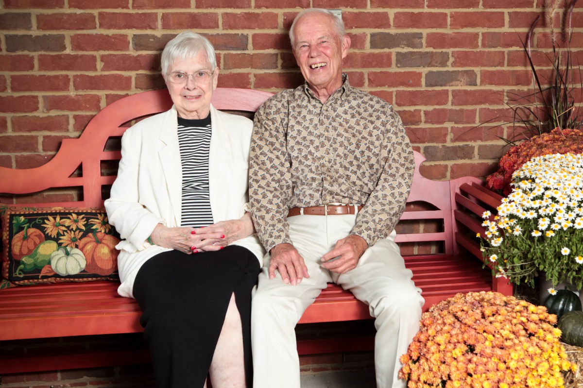 An elderly couple sitting together on a bench in front of a brick wall.
