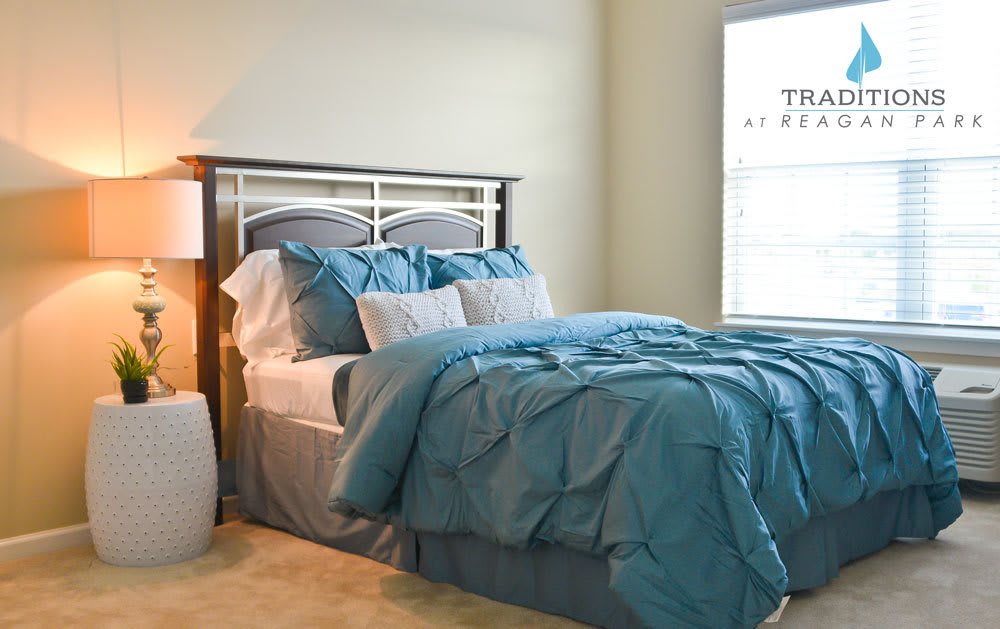 A cozy bedroom featuring a bed with blue bedding and pillows, a bedside lamp, and a window.