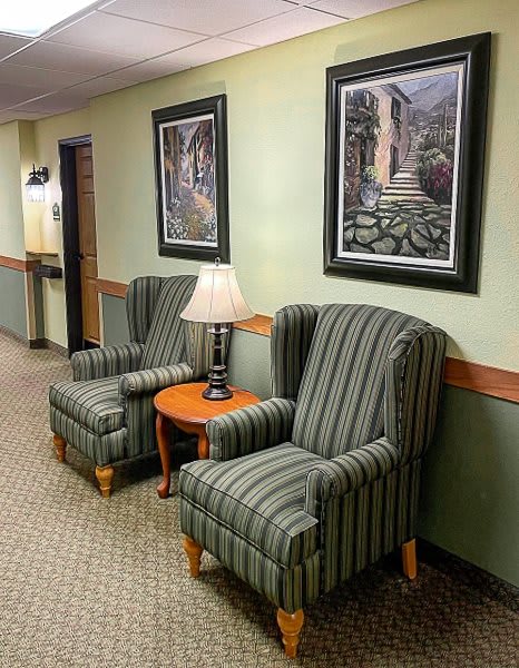 A hotel lobby area with two striped chairs, a small round table with a lamp, and framed paintings on the wall.