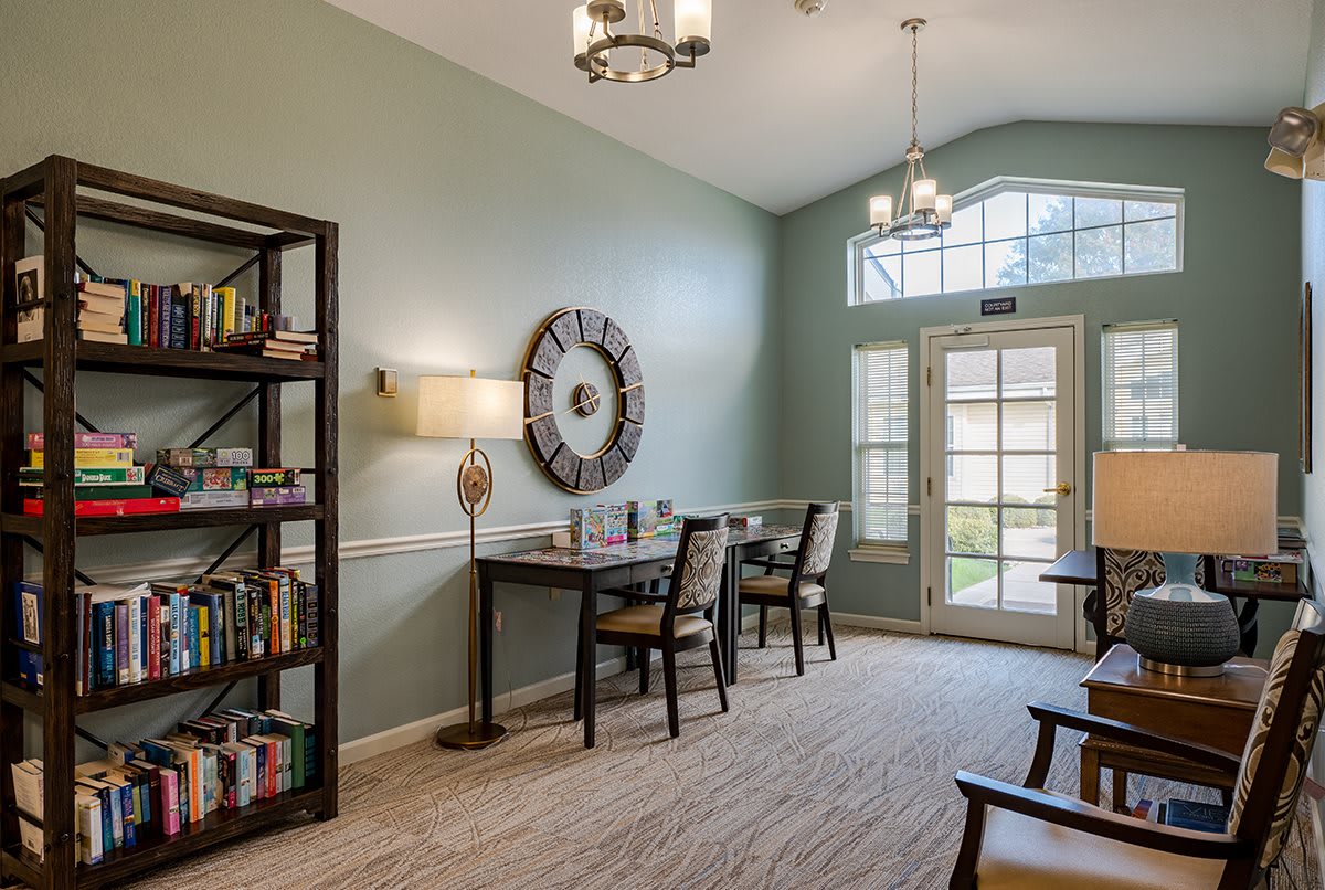 A cozy living room with bookshelves, a table with chairs, and large windows allowing natural light.