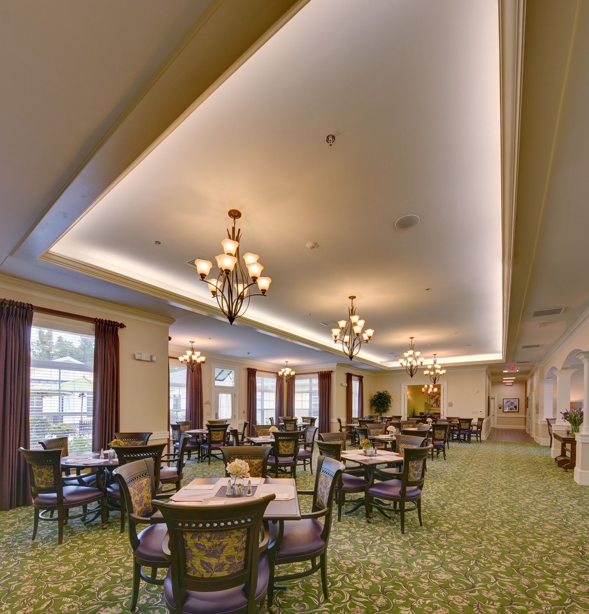 An elegant dining room with chandeliers, tables, and chairs arranged on a carpeted floor.