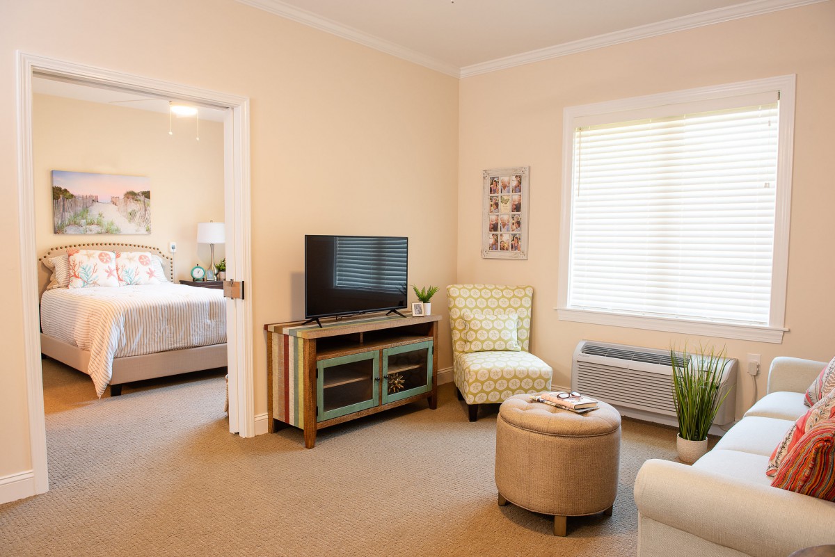 A cozy living room with a TV, chair, and window leading to a bedroom.