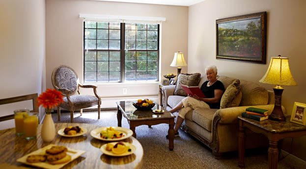 A cozy living room where a woman sits on a sofa, reading, with a window view and a table set with fruit.