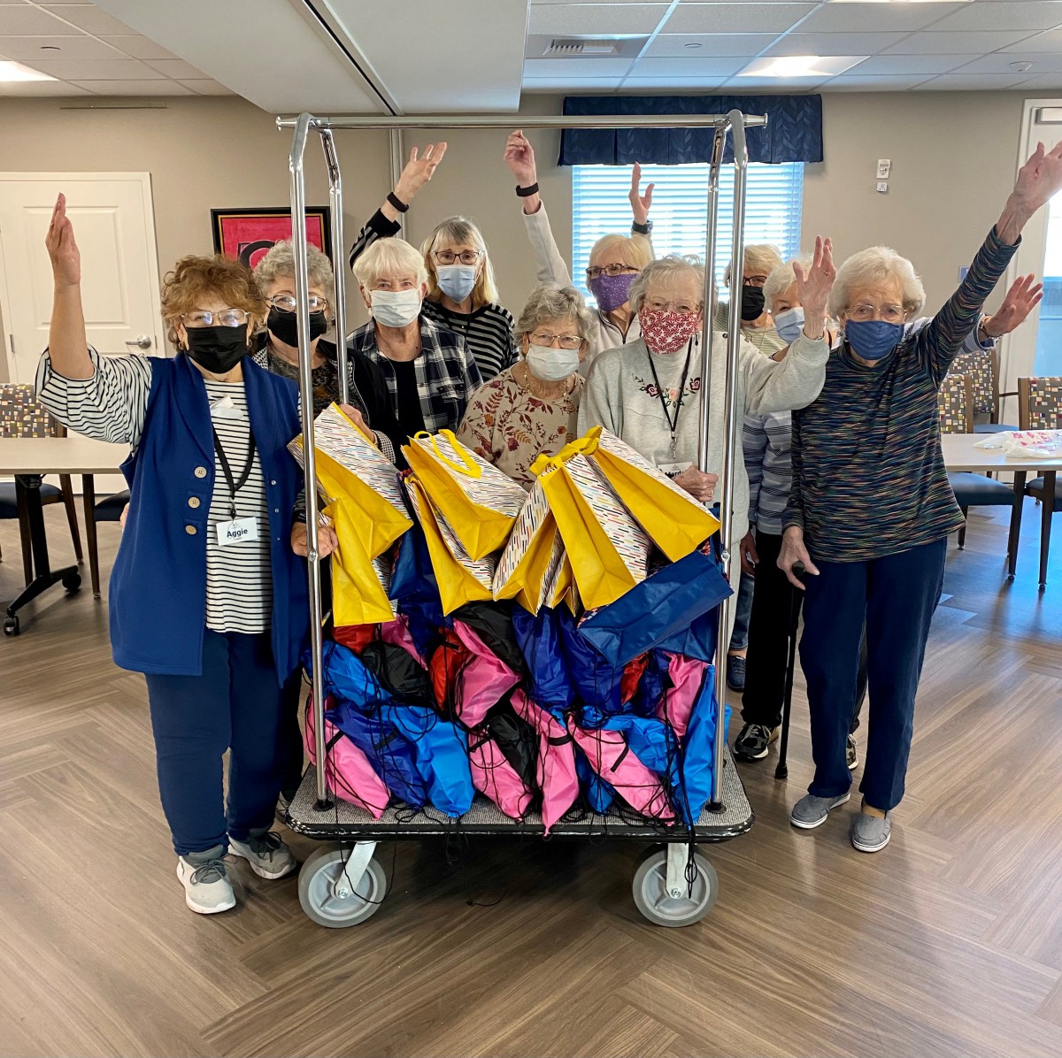 A group of seniors, wearing masks, enthusiastically gather around a cart filled with colorful bags in a community setting.