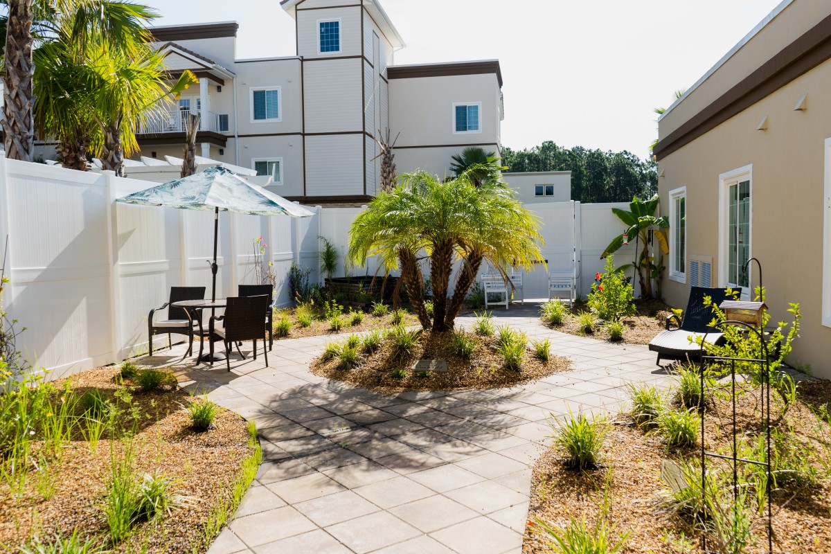 A well-landscaped courtyard with a central palm tree, outdoor seating, and a pathway under the bright sun.