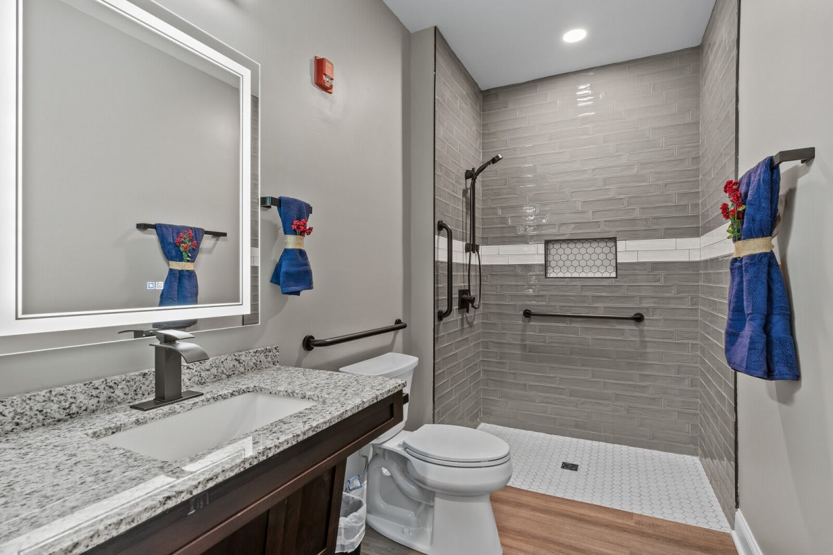 A modern bathroom with a spacious shower, sink, and toilet, featuring gray tile and accessible grab bars.