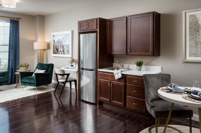 A modern apartment interior featuring a kitchen with dark wood cabinets and a stainless steel fridge, adjacent to a cozy living area with seating and a small dining table.