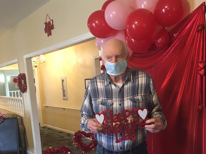 An elderly man wearing a mask holds a Valentine's Day decoration in front of festive balloons.
