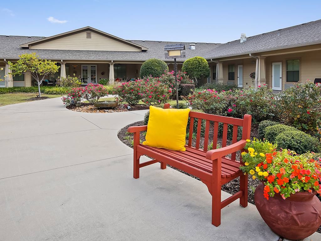 A peaceful outdoor scene with a red bench adorned with a yellow pillow, surrounded by a well-maintained garden and flowers, in front of a building.