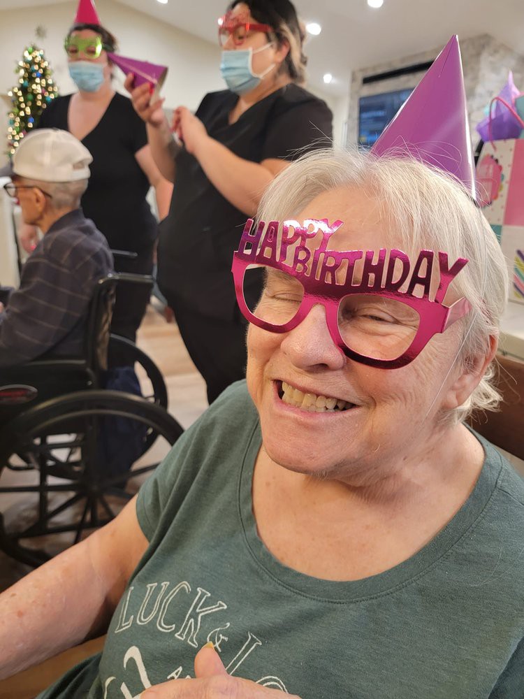 An elderly woman wearing 'Happy Birthday' glasses is smiling at a celebration.