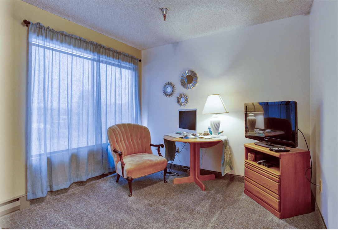 A cozy living room with a chair, desk, lamp, and television by a window with sheer curtains.