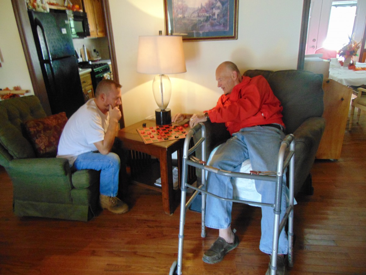 An elderly person and a younger man engage in a conversation in a cozy living room setting.