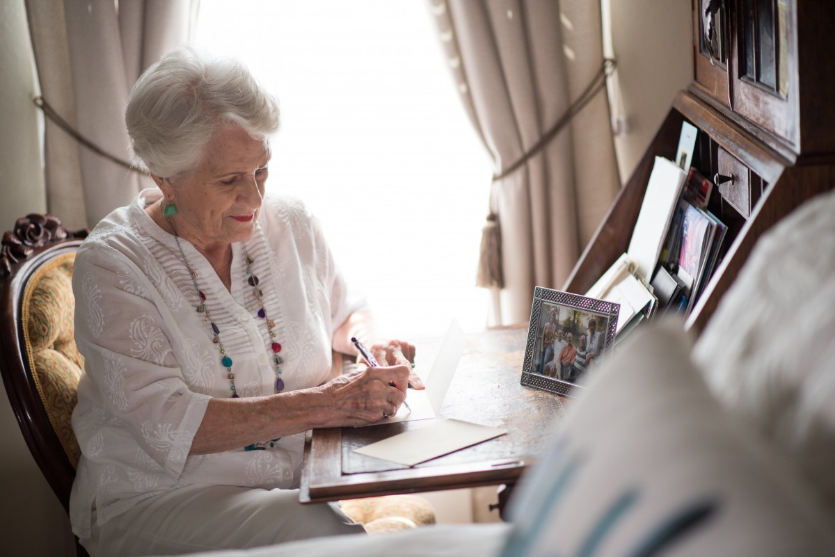 An elderly woman writing in a notebook at her desk.