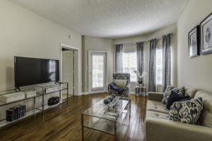 A modern living room with a TV, sofa, armchair, and a glass coffee table on wooden flooring.