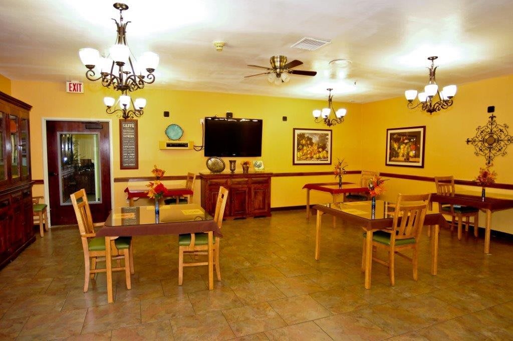 A cozy dining room with tables and chairs, featuring elegant chandeliers, a ceiling fan, wall-mounted television, and decorative artwork.