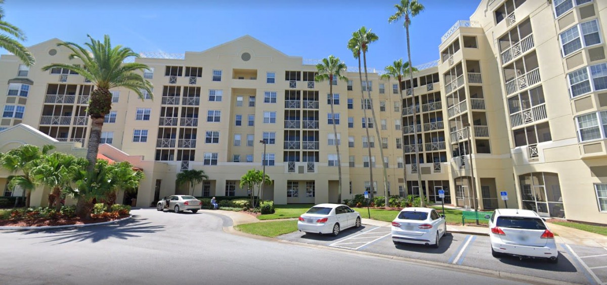 A residential apartment building with palm trees and a parking lot with parked cars.