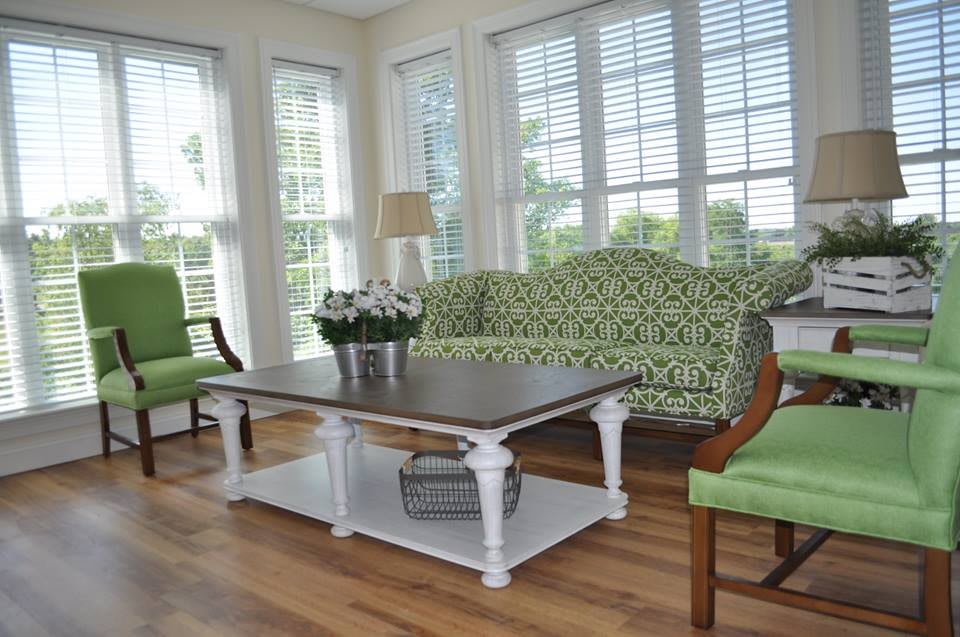 A bright living room featuring green furniture, a wooden coffee table, and large windows with blinds.