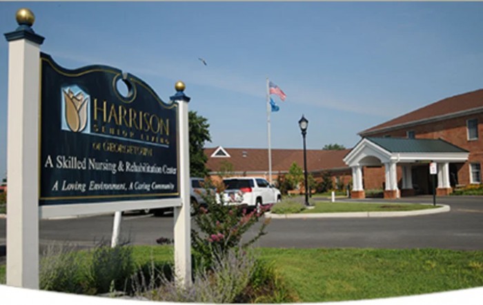 Entrance sign and view of the Harrison nursing and rehabilitation center with flags and parking area.