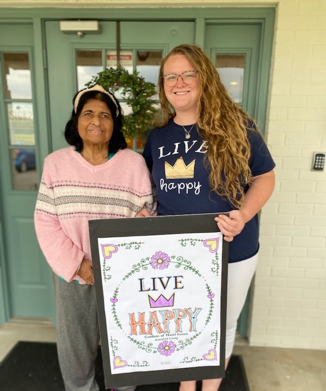 Two women smiling in front of a door, holding a sign that says 'Live Happy'.