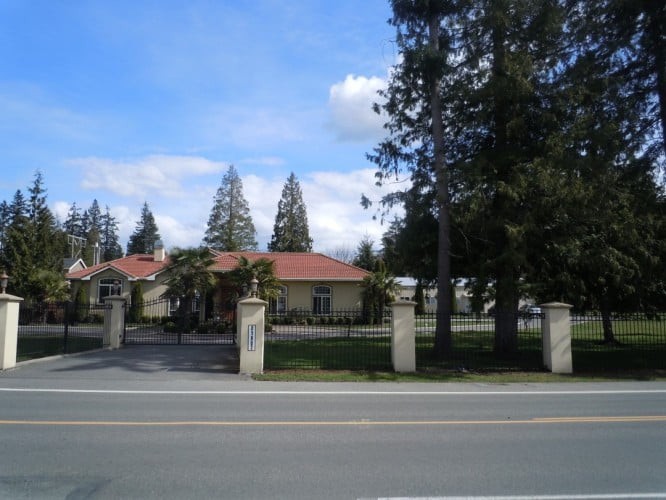 A suburban house with a red roof, surrounded by trees and a gated fence, viewed from across the road on a clear day.