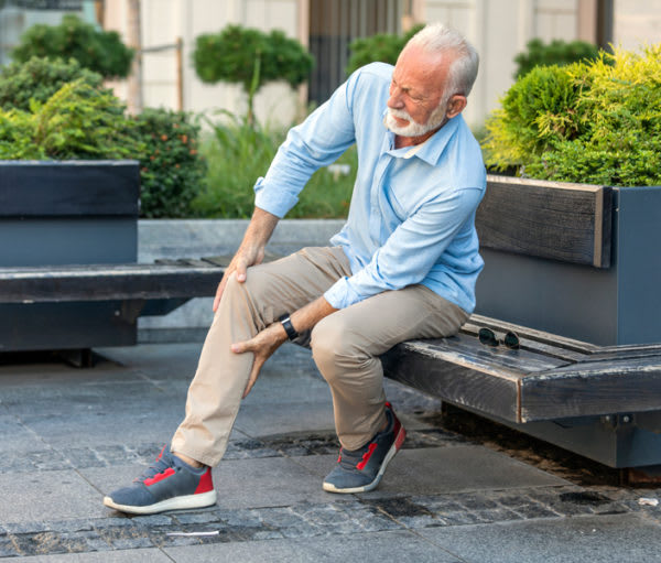 An elderly man sitting on a bench outdoors, holding his knee in discomfort.