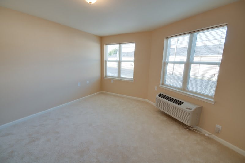 A bright, empty room with carpeted flooring and an air conditioning unit under the windows.