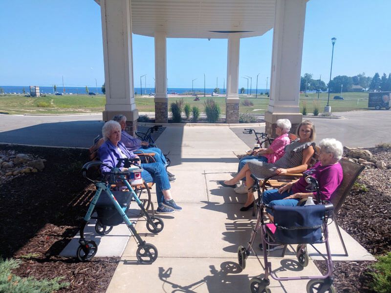 A group of seniors sitting outside on a porch, enjoying a sunny day with walkers beside them.