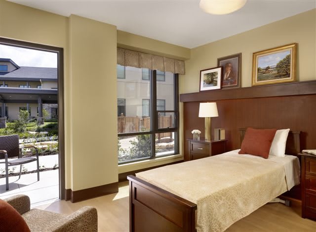 A cozy, well-lit bedroom featuring a single bed with wooden headboard, a bedside lamp, and large windows bringing in natural light.