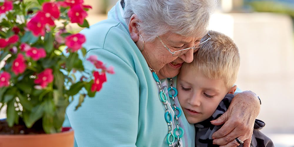 An elderly woman lovingly embraces a young child outdoors, surrounded by flowers.