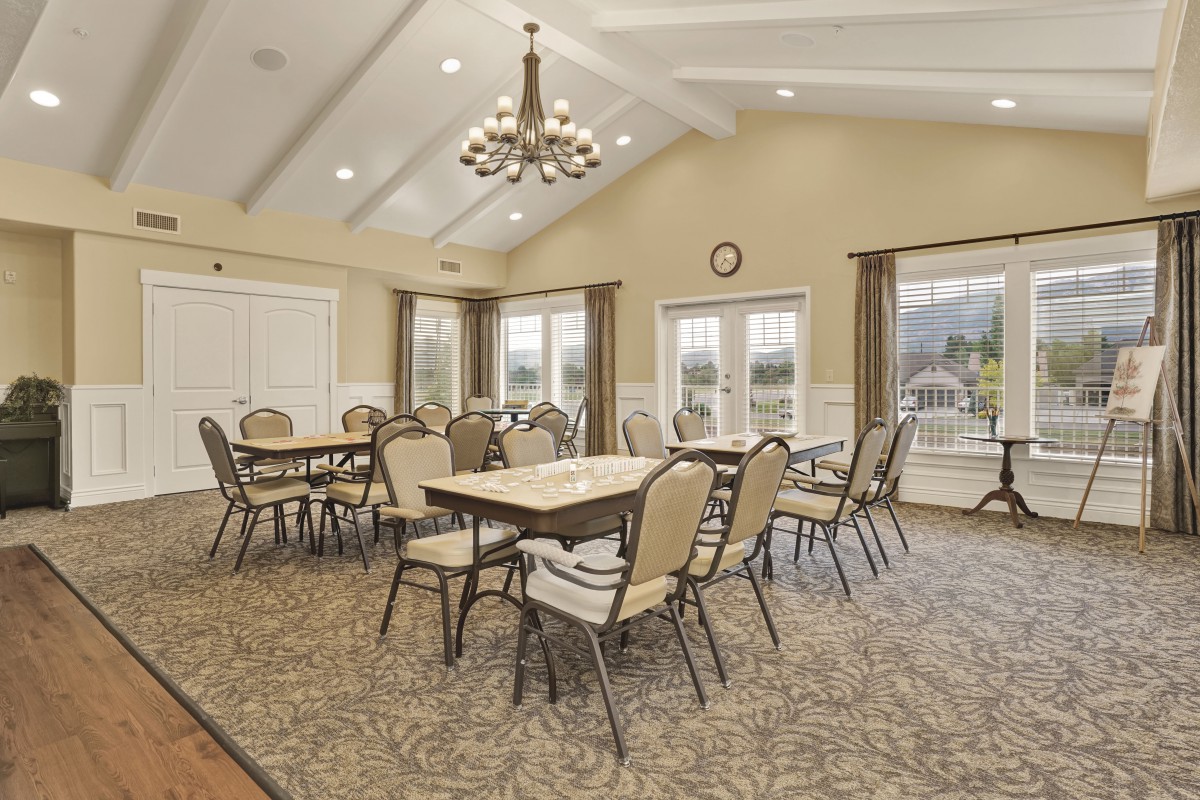 A spacious dining room with multiple tables and chairs, featuring a chandelier and large windows.