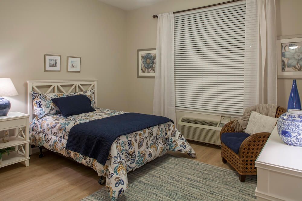 A cozy bedroom featuring a single bed with floral bedding, a comfortable chair, and large window blinds.
