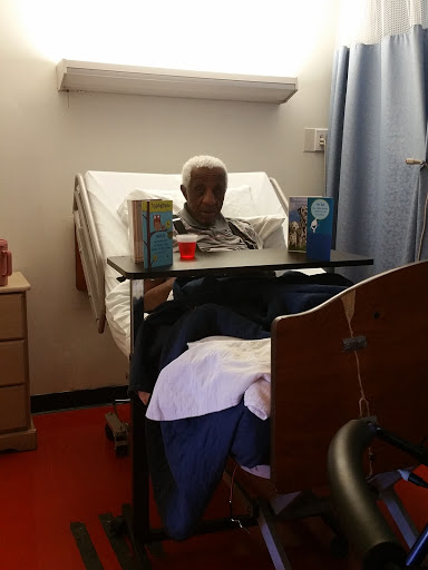 A senior patient sitting up in a hospital bed, surrounded by care items.